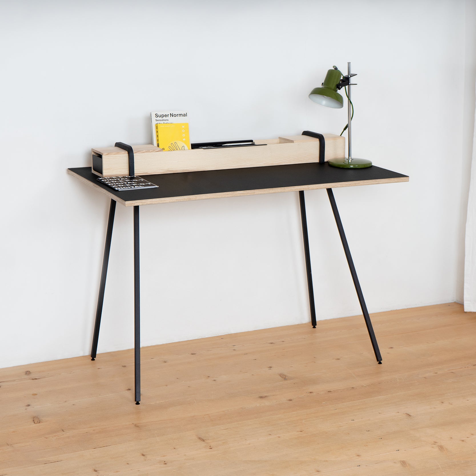 The Kurt by Moormann is a minimalist desk featuring thin black legs and a sleek black surface, complemented by a wooden organizer holding a magazine and yellow book. A small green lamp sits to the right. Positioned against a white wall on wooden flooring, it offers smart storage solutions.
