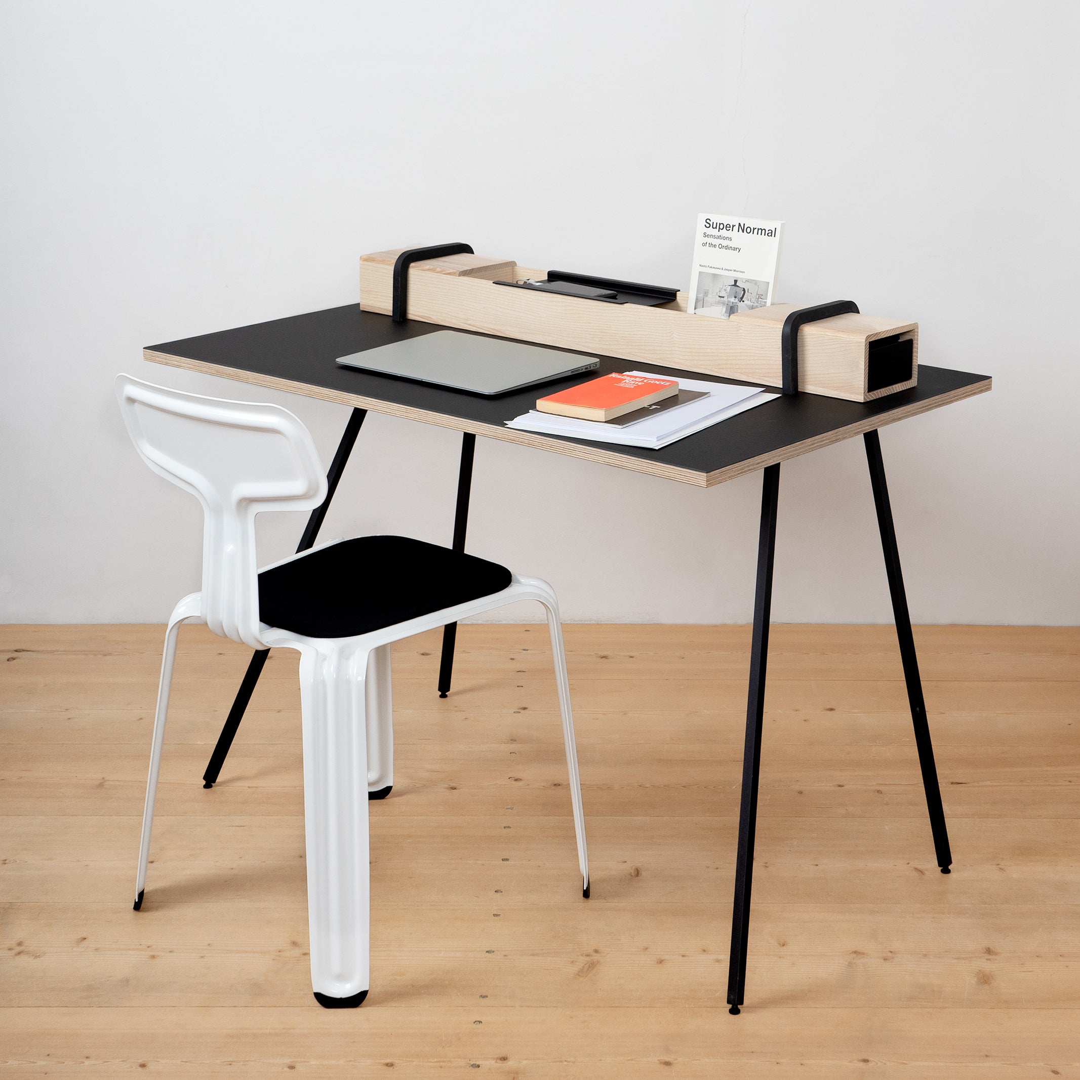 The Kurt desk by Moormann features a sleek black and wood finish with ample storage. It includes a white chair, wooden organizer, laptop, notebook, orange booklet, and Super Normal, all secured with a solid wood latch.