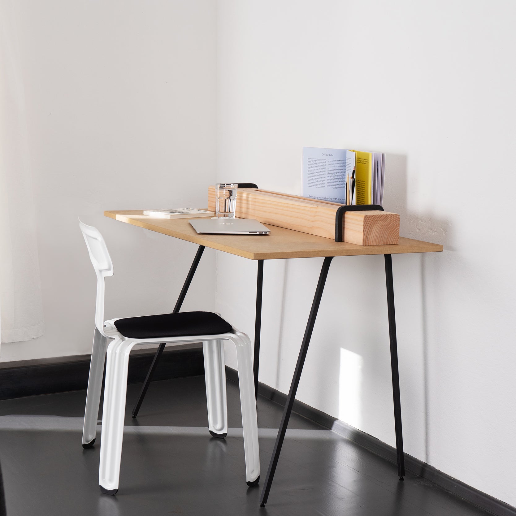 A minimalist setup showcases Moormanns Kurt desk with a wooden top on black metal legs, accompanied by a white chair. The desk holds books, a notepad, and a glass of water. Subtle storage complements the plain white wall in this secretary-inspired design.