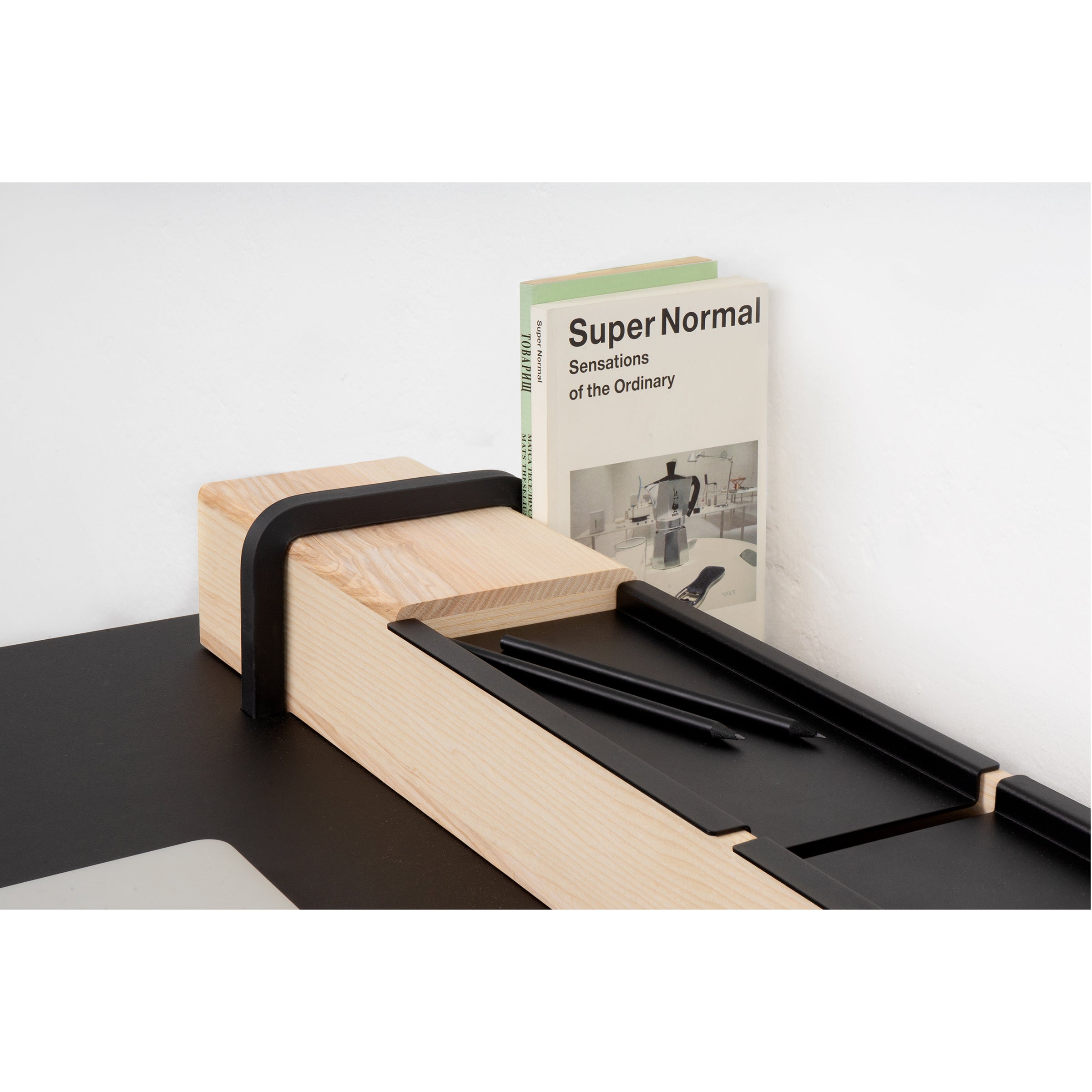 A sleek black and wood desk organizer from Moormann, similar to the Kurt model, holds Super Normal: Sensations of the Ordinary and two black pens. This minimalistic setup rests stylishly on a black surface against a white backdrop.