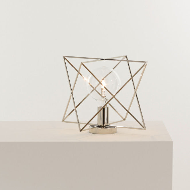 The KAIA LUM Table Lamp, currently on sale, features a star-shaped light encased in a metallic wireframe design with intersecting angular lines. This geometric lamp sits on a white surface, offering a modern and artistic contrast against its light-colored background.