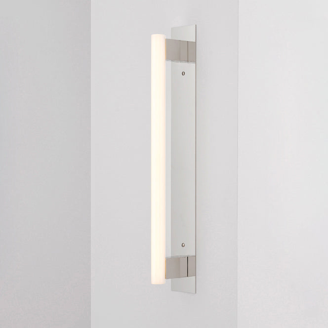 The MEA Plate by KAIA is a modern, wall-mounted vertical light fixture with a sleek, elongated design. It features a white light and brushed brass accents on a pale gray wall, offering an aesthetic and contemporary look.
