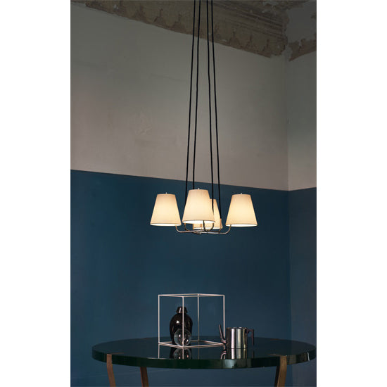 A modern pendant light from Kalmars Corona collection, accented with polished brass and elegant silk shades, hangs above a round glass table featuring a decorative metallic cube frame and a sleek black bowl. The wall is half-painted in teal and cream, while the textured ceiling is partially visible.