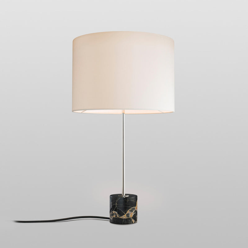 The Kalmar Kilo Nero Portoro Marble Table Lamp features a sleek cylindrical white shade and thin metal stem. Its Nero Portoro marble base with subtle veins enhances the visible black power cord against a light gray background.