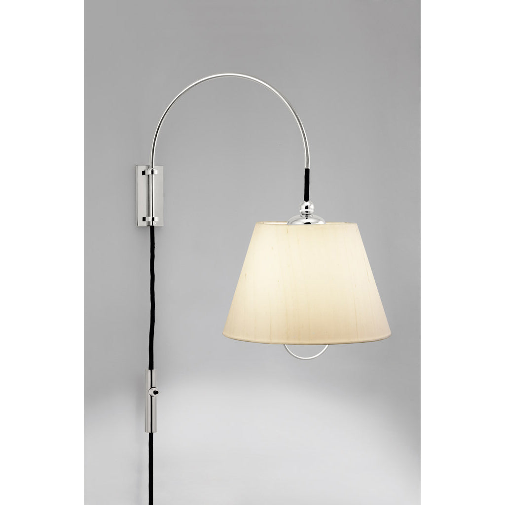 A modern wall lamp, the Kalmar Reibe Wall, features a sleek metal arc and light beige fabric shade on a gray wall. Enhanced by a black cord, this polished brass fixture serves as the ideal minimalist bedside companion.