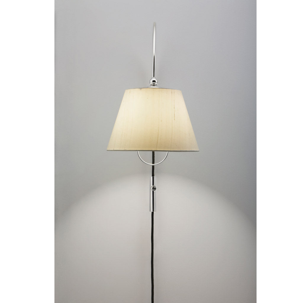The Kalmar Reibe Wall offers a modern wall-mounted luminaire with a cream conical lampshade and polished brass stand, casting soft light on a gray background, making it an ideal bedside companion.