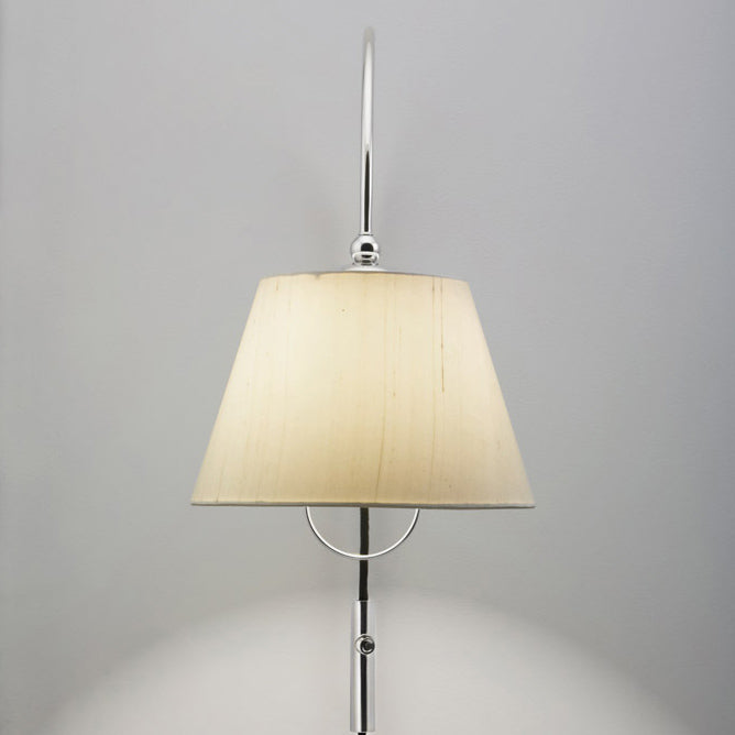 The Kalmar Reibe Wall luminaire showcases a modern design with a curved silver arm and cream cone-shaped lampshade. Its polished brass details provide elegant bedside lighting, casting a subtle glow against the light gray wall.