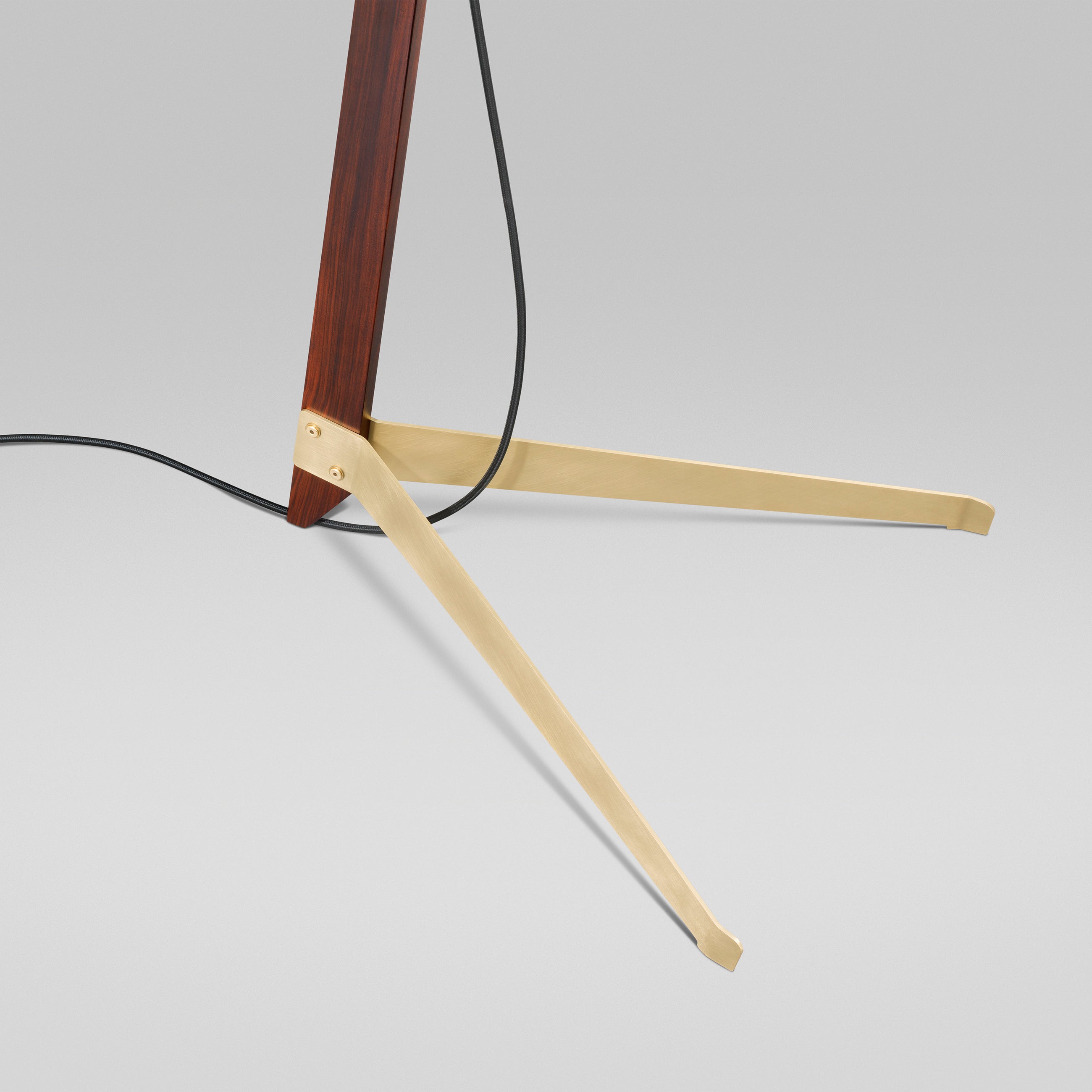 Close-up of the Billy Floor Ilse Crawford Edition by Kalmar, showcasing a modern tripod stand with two satin-brass feet and a dark rosewood stem, featuring a black cord hanging down against a plain light gray background.
