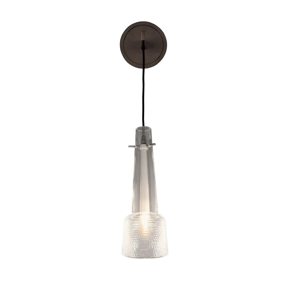 The Kalmar Keule Wall features a modern pendant luminaire with a tapered clear glass shade and textured design. It hangs from a black cord attached to a black bronze ceiling mount. The bulb provides a subtle glow, accentuating the glass details for an elegant, minimalist appearance.