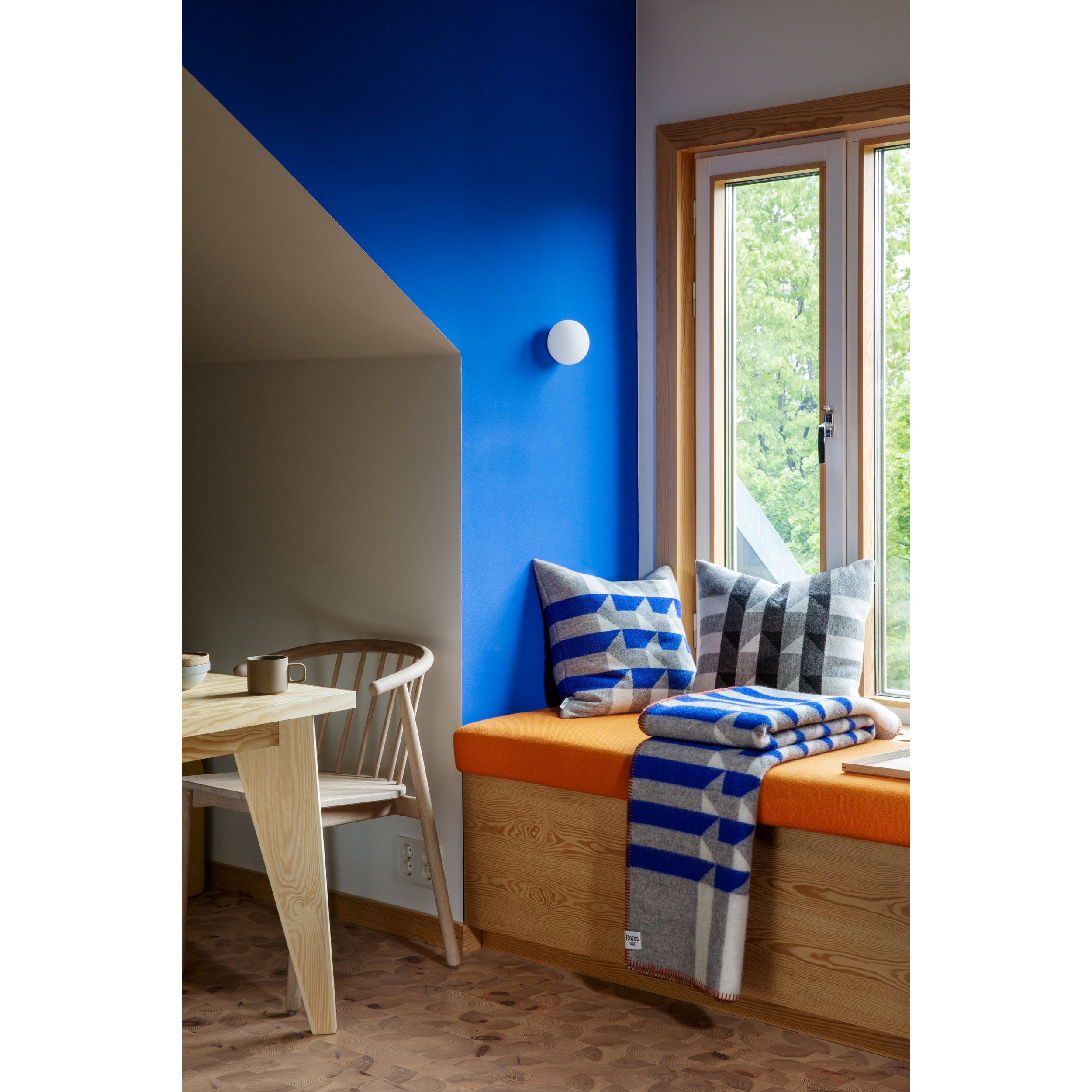 In a cozy nook, an orange cushioned seat by the window showcases Røros Kvam Cushions and wool blankets in blue and gray stripes. A nearby wooden chair and table complement the setup, while a bright blue wall contrasts against the lush greenery visible outside.