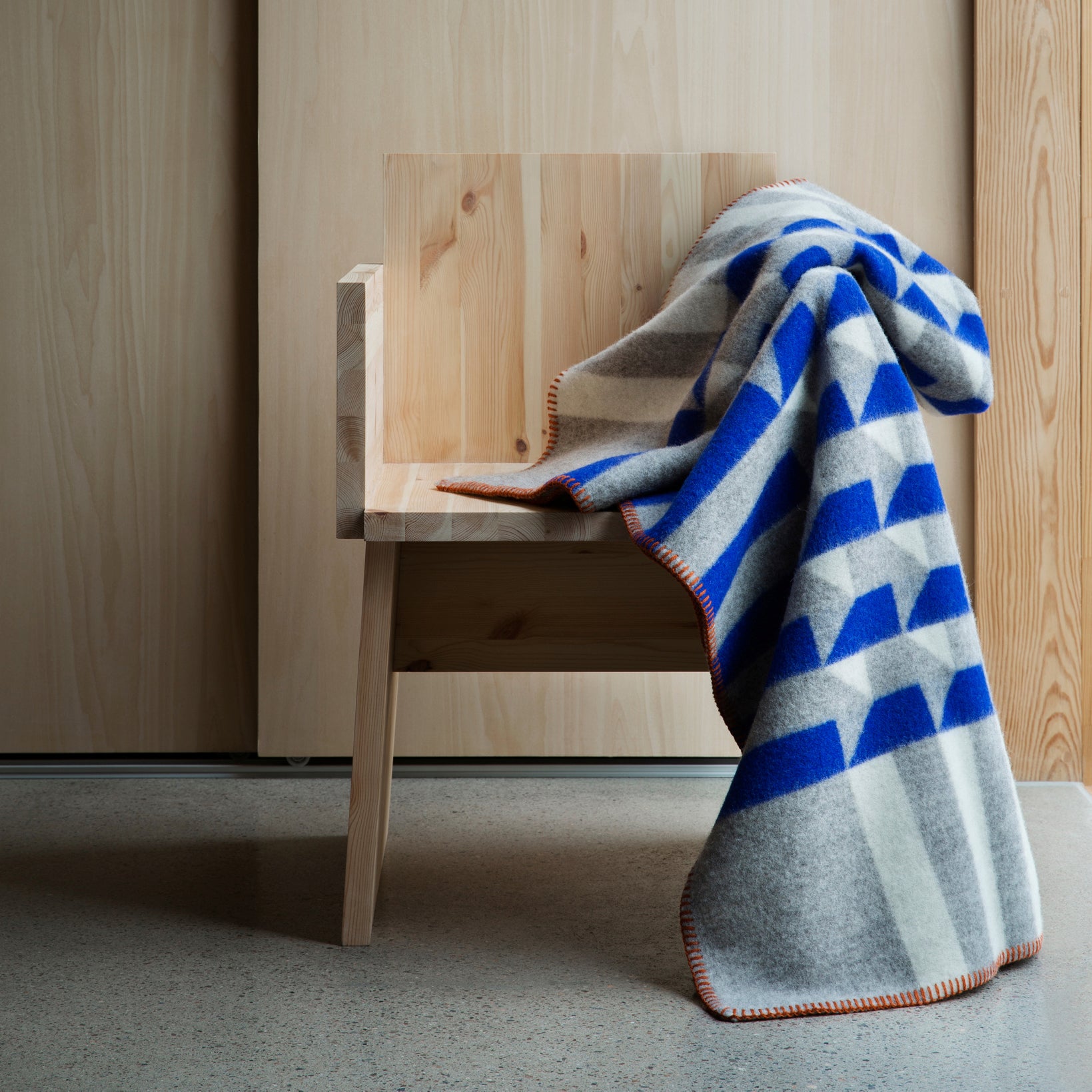 The Kvam Large Throw by Roros, made from soft Norwegian lambswool, features blue, gray, and white jacquard weave triangles with an orange-trimmed edge. It sits on a wooden chair, enhancing the minimalistic ambiance of a smooth floor and wooden backdrop.