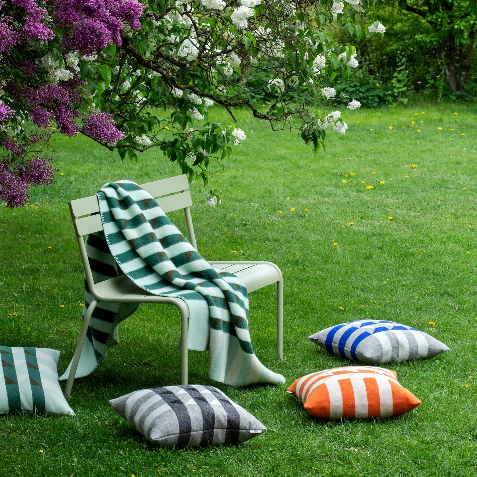 A green chair set on grass is draped with a striped Røros Tweed blanket made from Norwegian lambswool, under a blossoming tree with white and purple flowers. Surrounding it are Kvam cushions by Roros in various striped colors like green, black, blue, and orange.