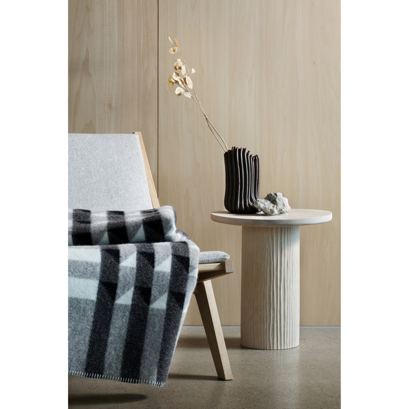 In a minimalist room, a light-colored wooden chair adorned with the Roros Kvam Large Throw in gray and black stands beside a round wooden table holding a black vase with dried branches and a small decorative object, all set against light wood paneling.