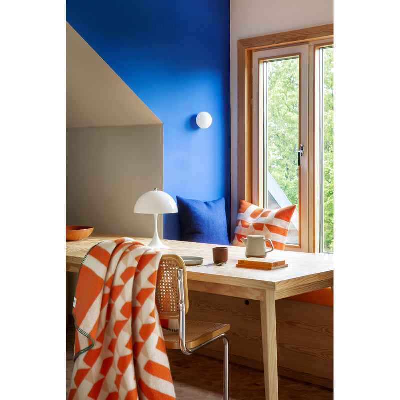 A bright room showcases a wooden desk with the Roros Kvam Large Throw draped over a chair. A white lamp, teacup, and stack of books rest on the desk. A blue wall features a window offering a lush green view beside a cushion with blue and orange patterns.