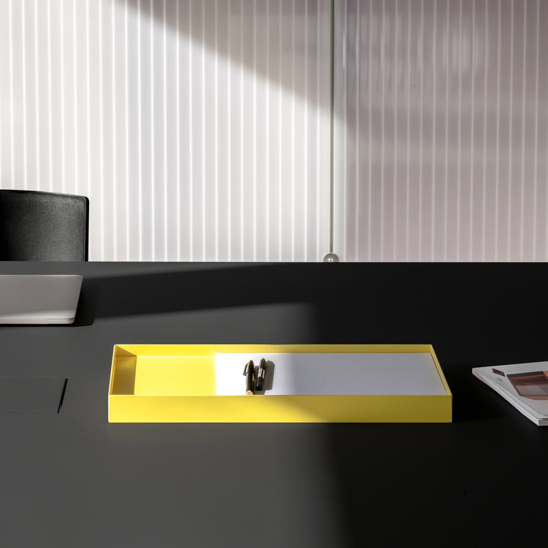 The minimalist L3 COURT desk by Loehr features a closed laptop on the left and a yellow steel tray with an object at the center. A magazine peeks from the right, as soft lighting casts artistic shadows on the corrugated backdrop, enhancing this serene setting.