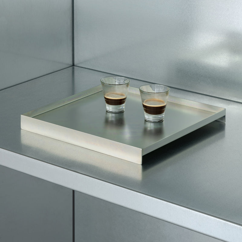 Two small glasses of espresso rest on the L3 COURT tray by Loehr, elegantly placed on a reflective metallic kitchen counter. The minimalist setting features clean lines and a monochromatic palette, exuding modern sophistication.