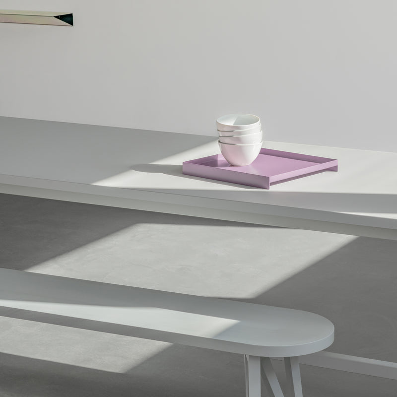 In a minimalist scene, the Loehr L3 COURT long white table and bench pair features prominently. A stack of white bowls sits on a pastel pink serving tray, casting soft shadows. Sunlight introduces geometric patterns on the plain backdrop, enhancing the artistic vibe with simplicity and elegance.