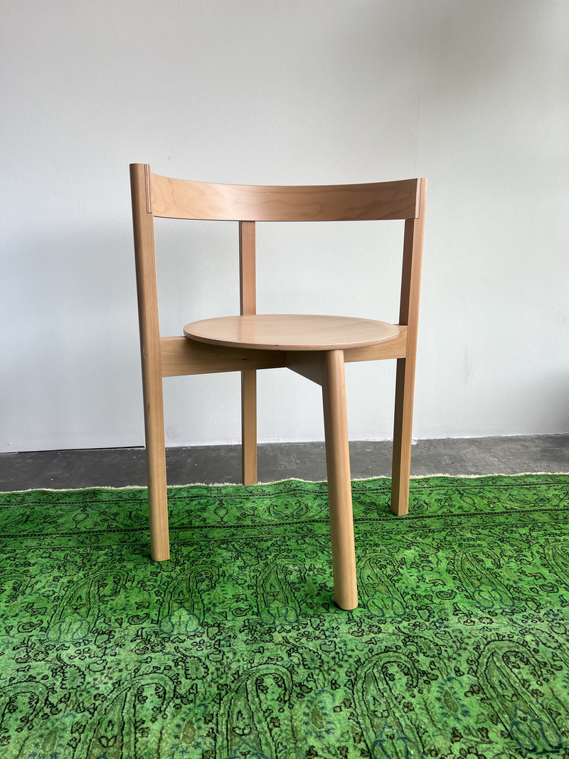 The L5 JAZZ Armchair by Loehr, with its minimalist design of a wooden round seat and three legs, alongside a curved plywood backrest, rests on a vibrant green patterned rug against a plain white wall and gray floor.