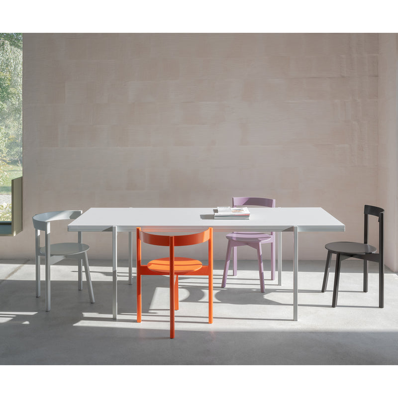A modern dining room has a white table surrounded by five L5 JAZZ Armchairs from Loehr, each in gray, orange, purple, black, and white. The solid wood chairs are beautifully highlighted as sunlight streams through the window, casting soft shadows on the floor.