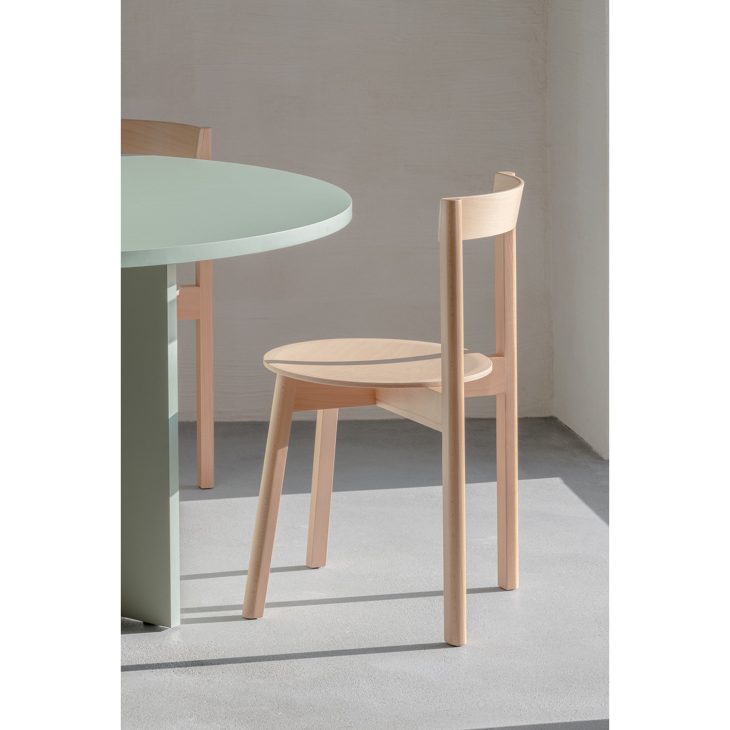 A minimalistic setting features a Loehr L5 JAZZ Side Chair with a round seat, paired with a round light green table, casting iconic silhouettes and soft natural shadows on the light gray floor.