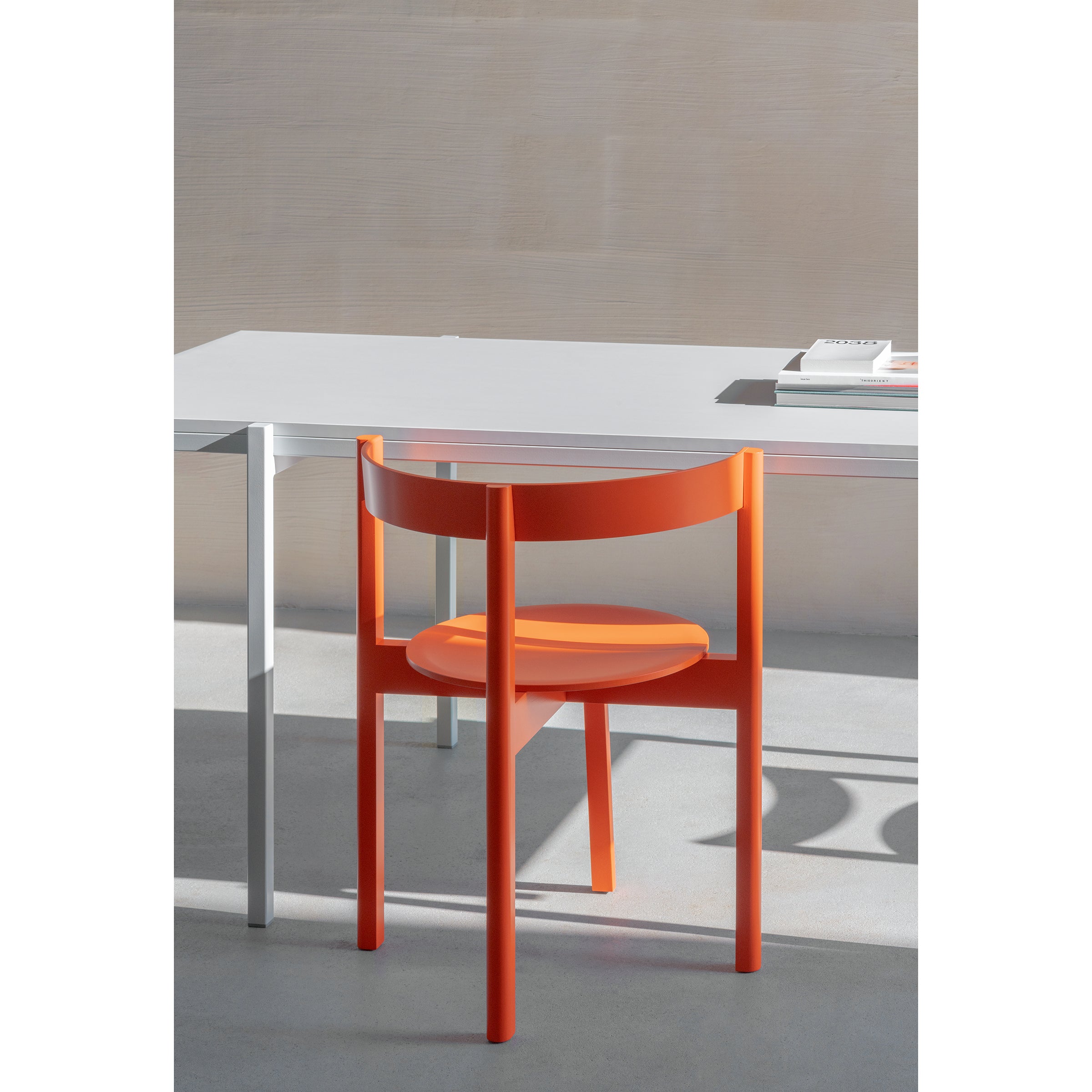 In a minimalist room, a white desk pairs with the L5 JAZZ Armchair by Loehr. Sunlight casts gentle shadows on the light gray floor, enhancing the serene ambiance. A few white books on the desk contribute to the clean and modern atmosphere.