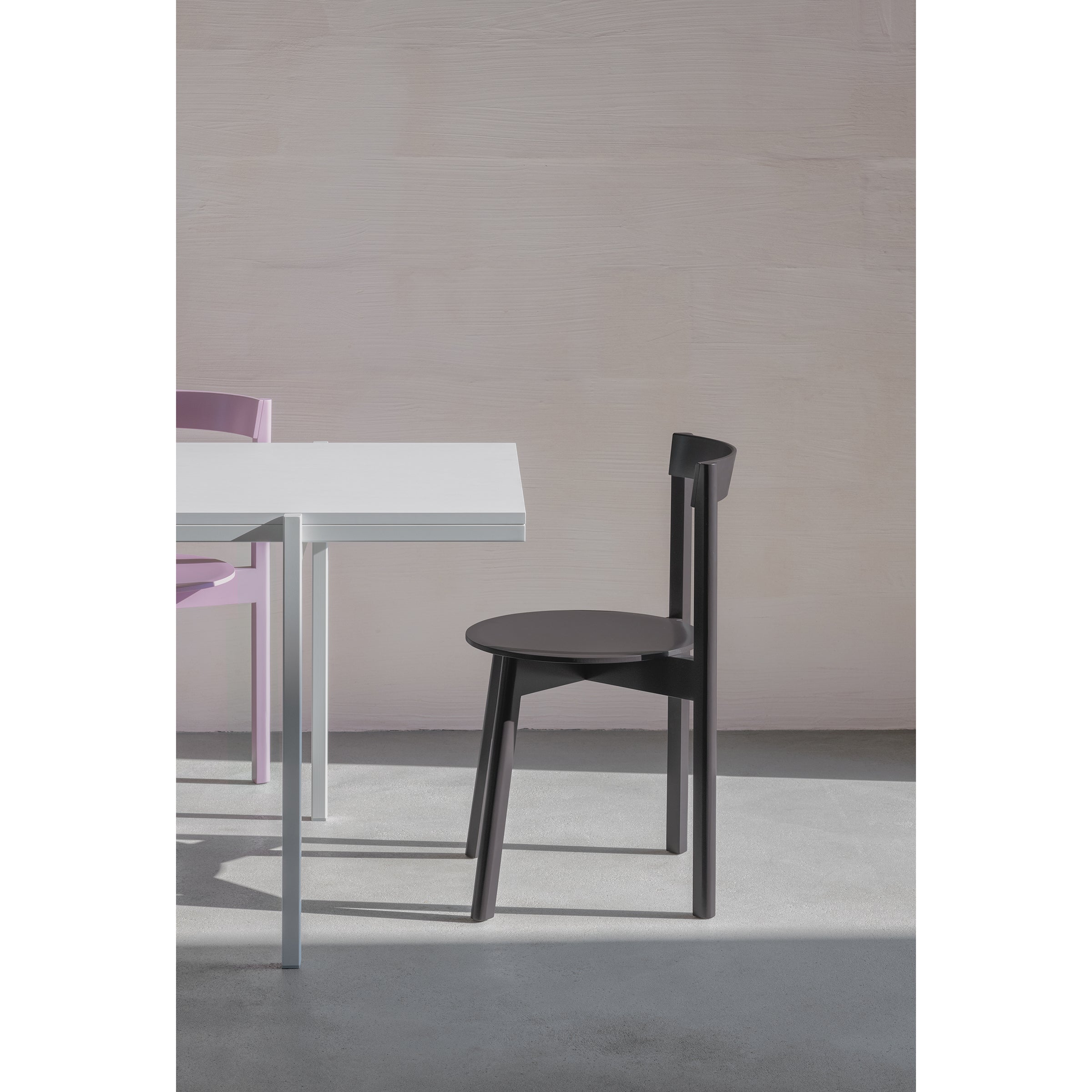 A minimalist room features a white table and the sleek silhouette of Loehrs black L5 JAZZ Side Chair on a smooth, light-colored floor. A touch of a purple chair adds depth against plain walls that accentuate the clean aesthetic.