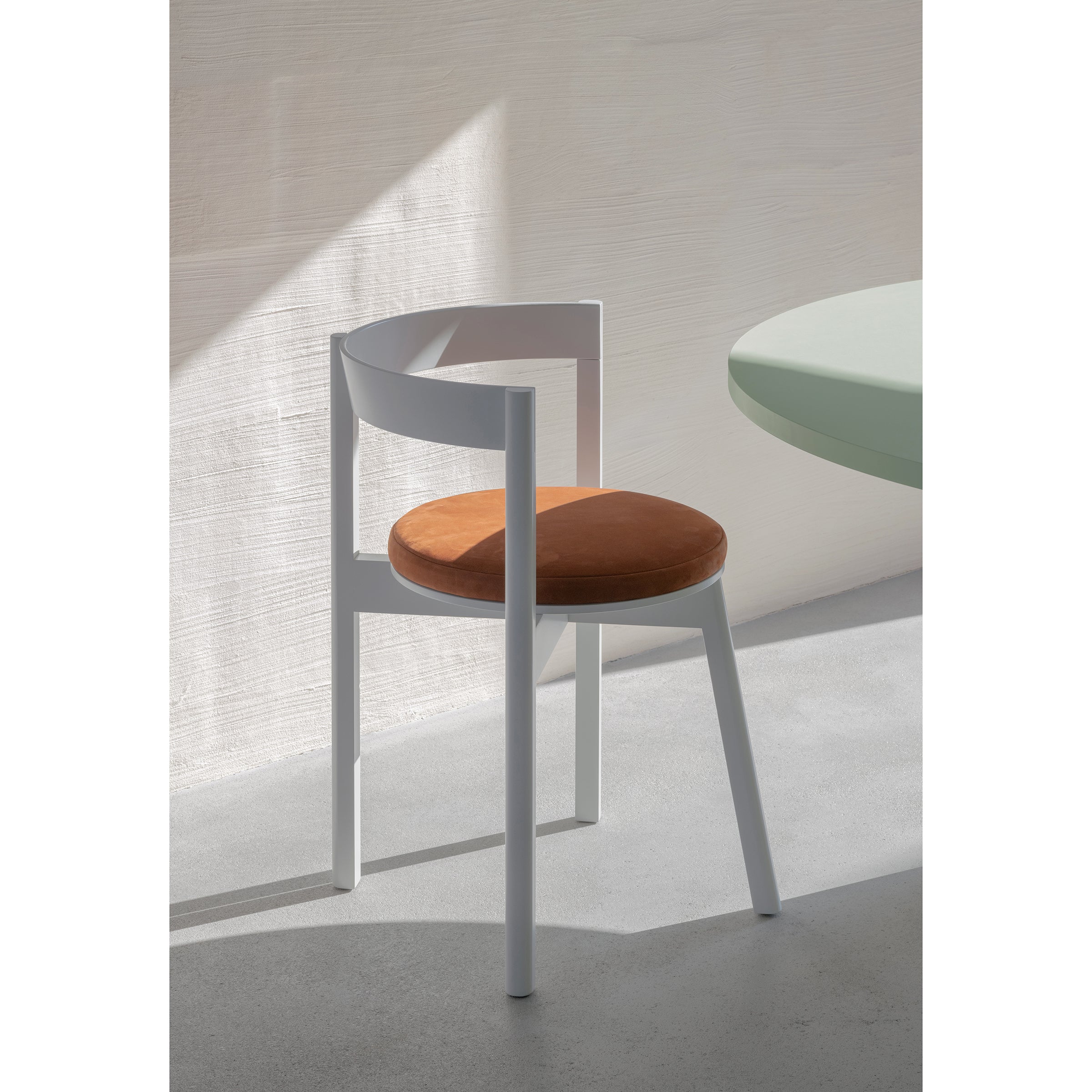 The L5 JAZZ Armchair by Loehr, showcasing a minimalist design with a white curved frame and orange cushioned seat, complements the light green solid wood dining table. Soft sunlight casts shadows on the light gray floor and textured white wall, enhancing the serene atmosphere.
