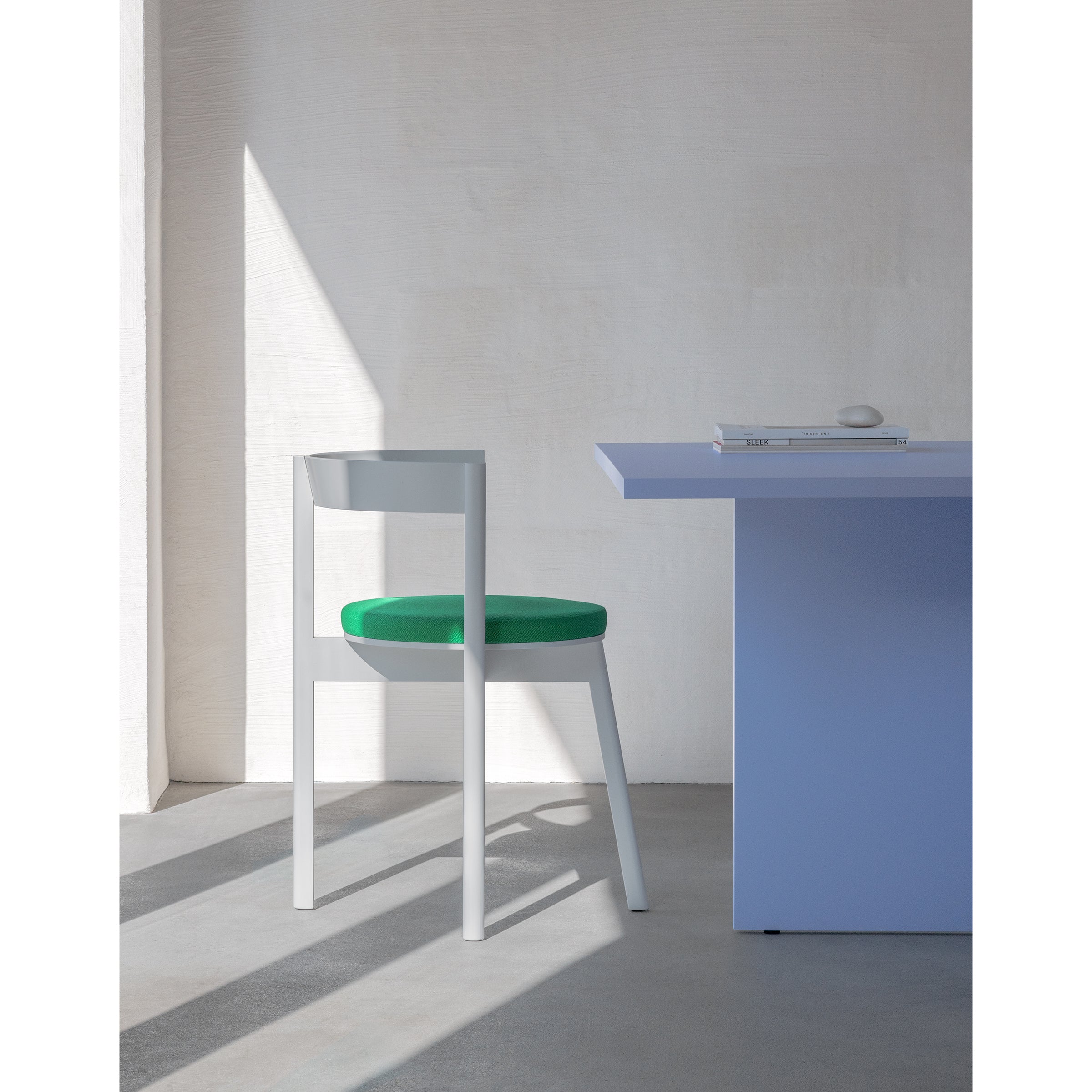 A minimalist room features a Loehr L5 JAZZ Side Chair with a green cushion next to a blue table. Sunlight casts angular shadows on the white wall and floor, creating a serene atmosphere. Its iconic silhouette complements the sparse items on the table.
