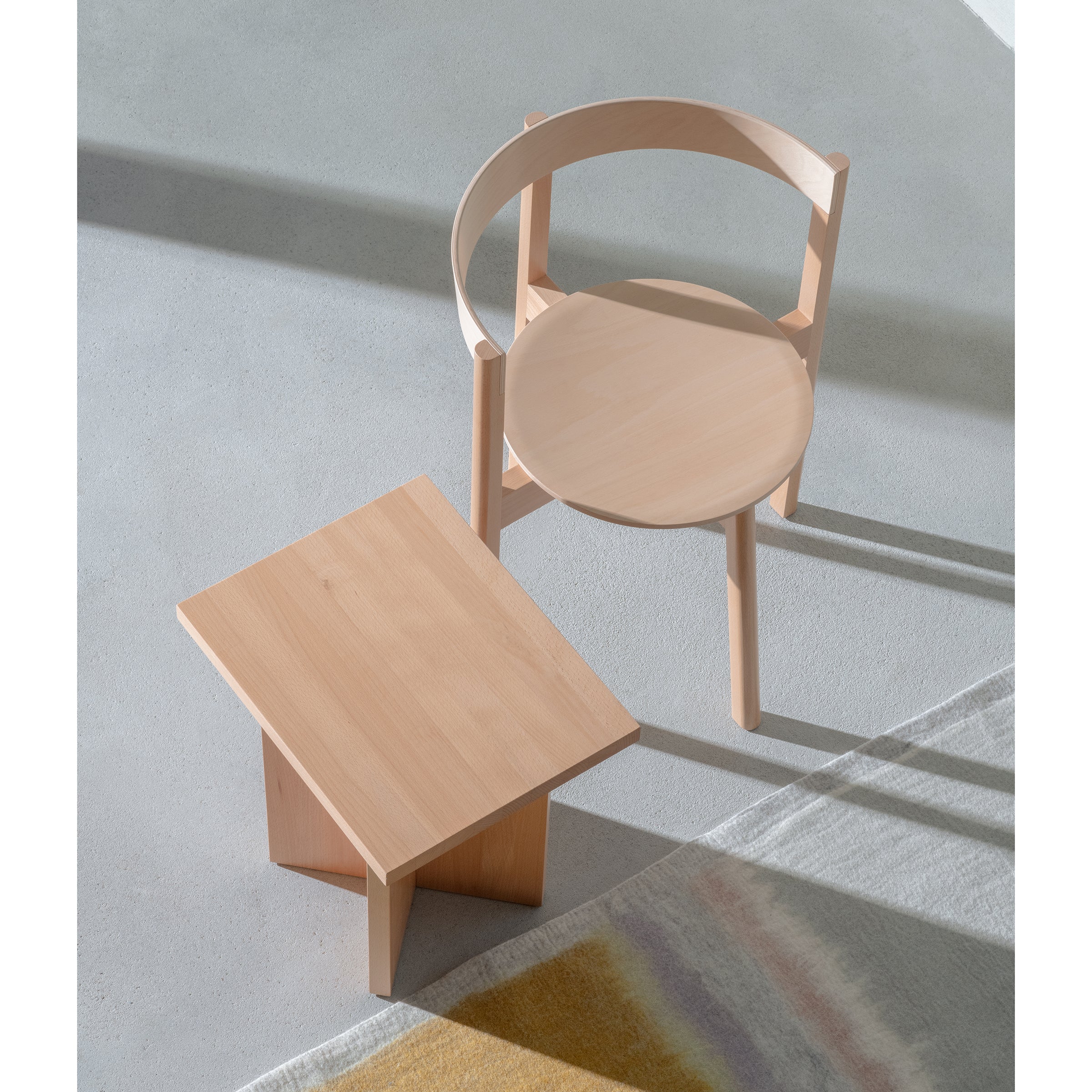 The L5 JAZZ Armchair by Loehr, featuring a plywood backrest, sits next to a rectangular wooden side table on a light-colored floor. Sunlight partially illuminates them while a patterned rug is seen in the bottom right corner.