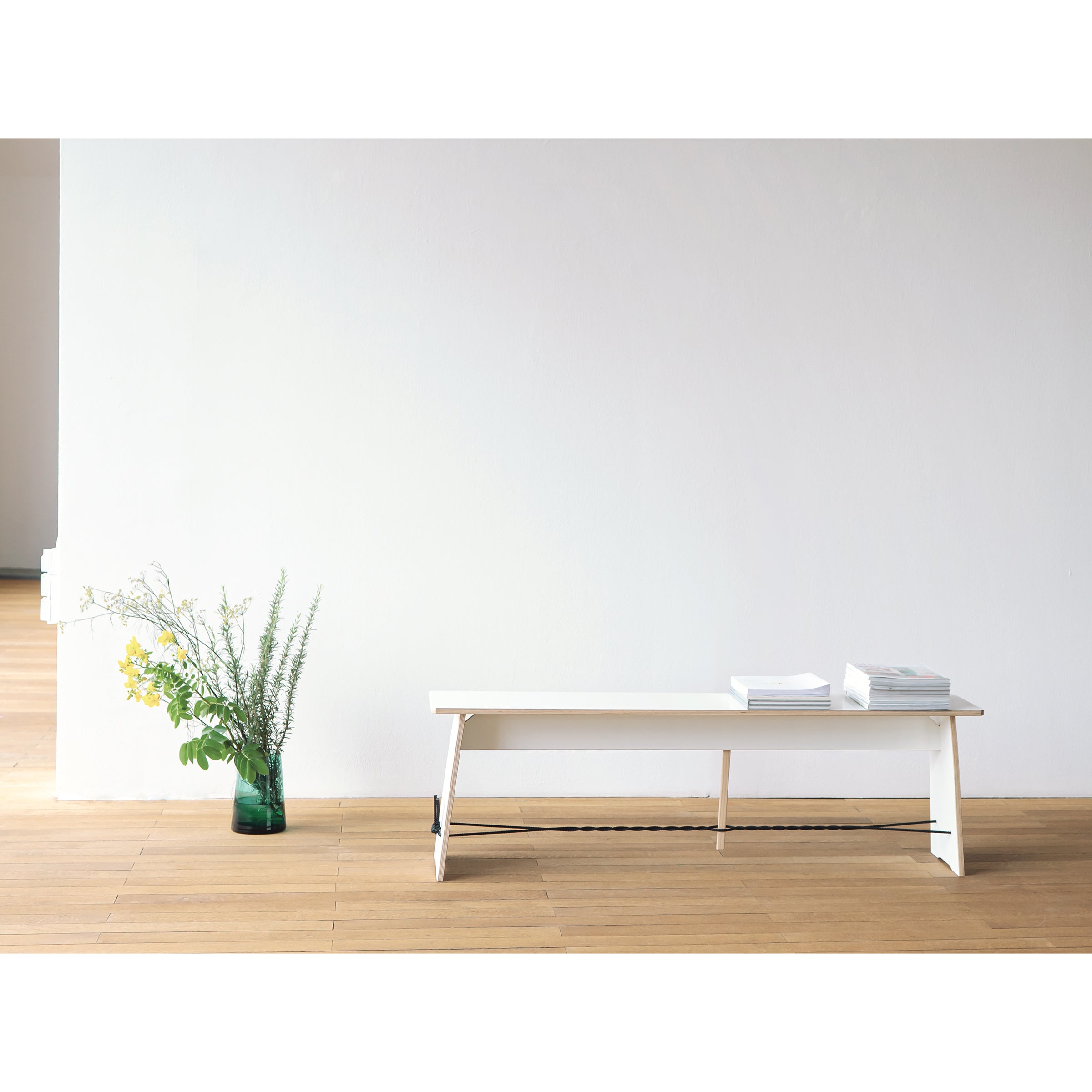 A minimalist room with wooden floors features designer furniture, including Moormanns Langer Max bench adorned with books on one end. Nearby, a green glass vase holds yellow and green flowers. The plain white walls enhance the well-lit, uncluttered atmosphere.