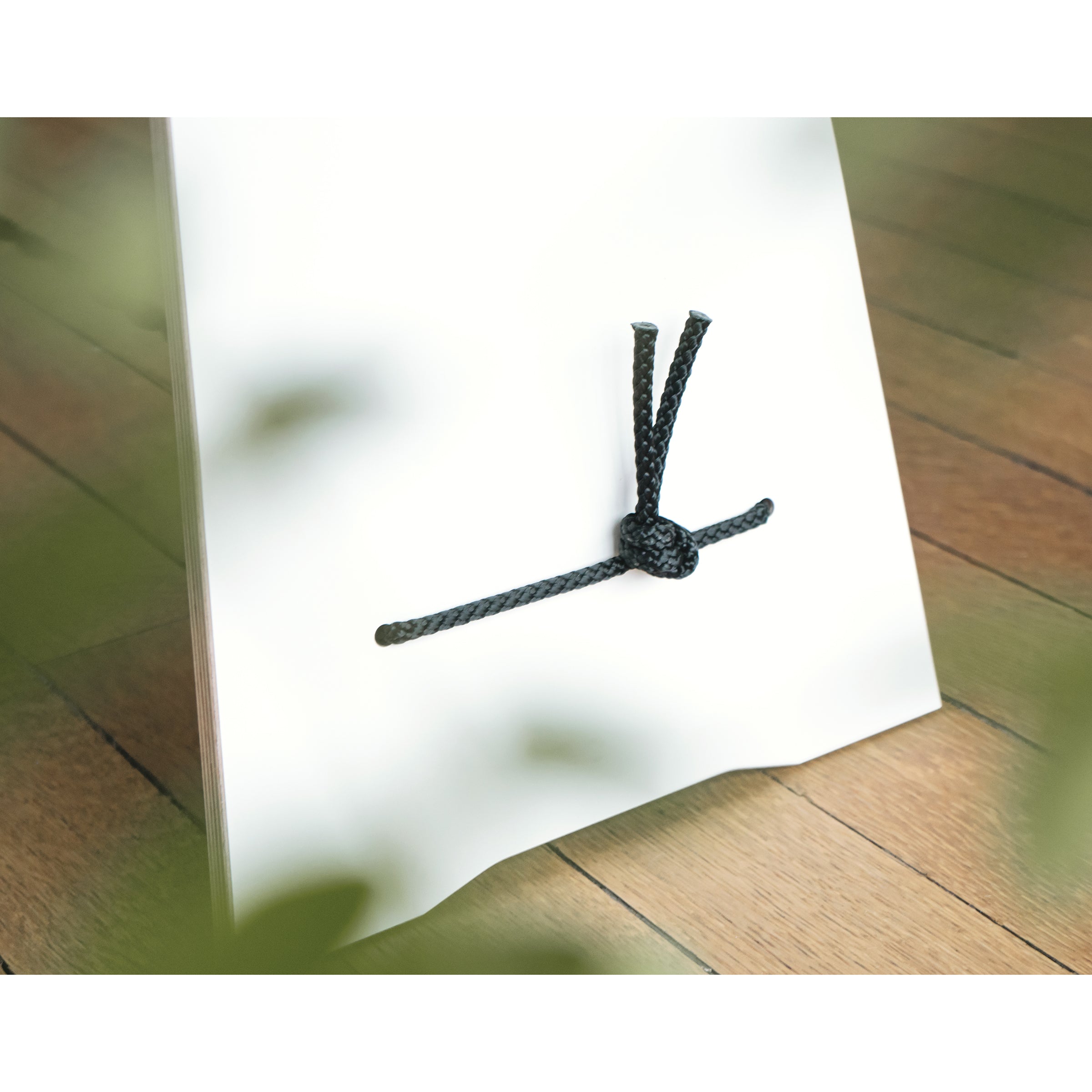 The Langer Max by Moormann features a white surface adorned with a black rope tied in a decorative knot, echoing designer furniture. The rope threads through the surface, with loose ends extending upwards against a wooden floor background and blurred green leaves in the foreground.