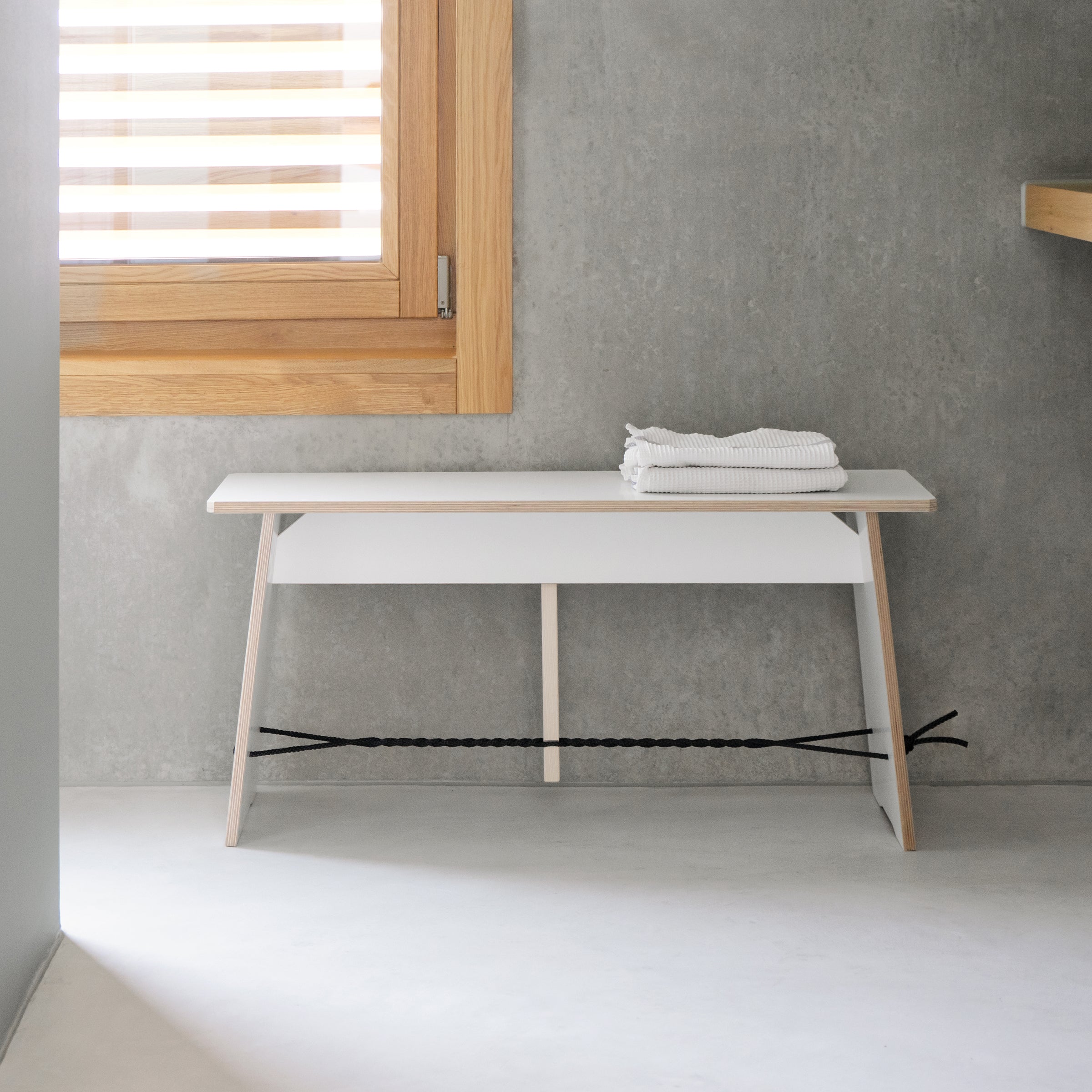 A minimalist bathroom with concrete wall and floor showcases designer furniture, including Moormann’s Langer Max—a small white bench under a wooden-framed window. A folded towel lies on the bench, supported by a sleek black diagonal design beneath.