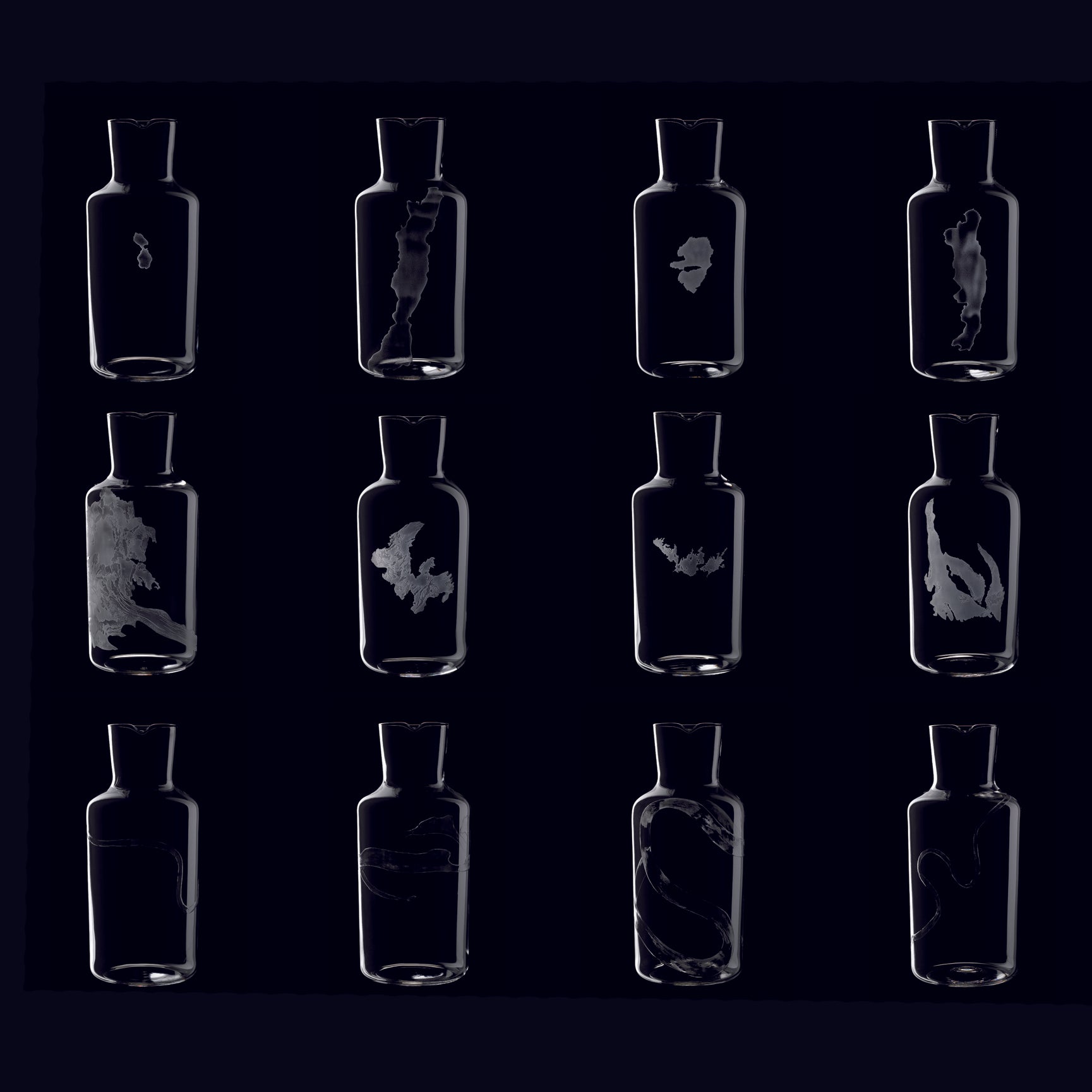 The Lobmeyr Fortune Series features twelve mouth-blown crystal bottles arranged in a grid on a dark background, each showcasing unique, abstract white forms resembling smoke or ink in water, offering an exquisite display of fluid elegance and craftsmanship.