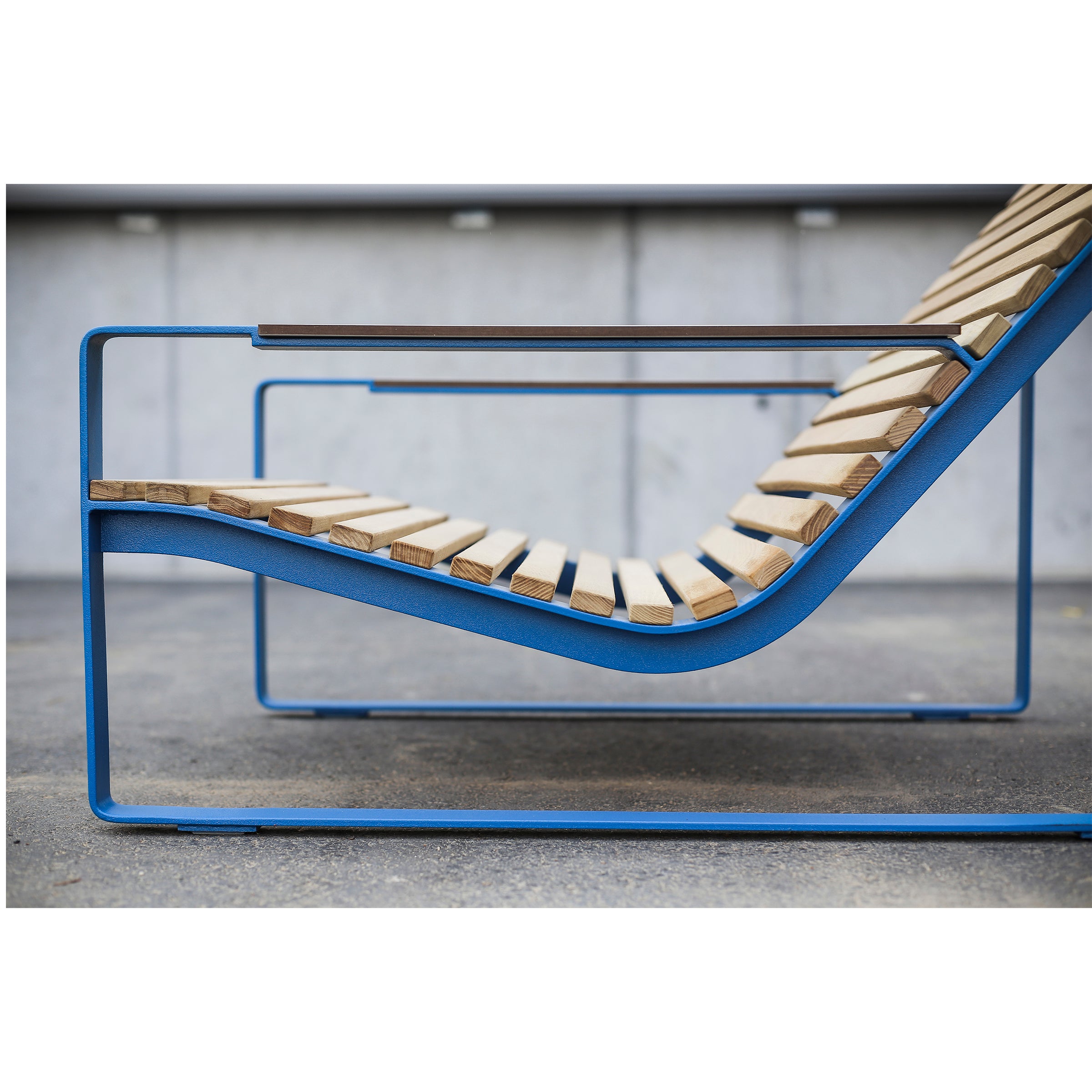 The Preva Lounger by Egoe showcases a sleek design with a blue metal frame and wooden slats against a concrete backdrop, emphasizing its minimalist aesthetic and high-quality materials.