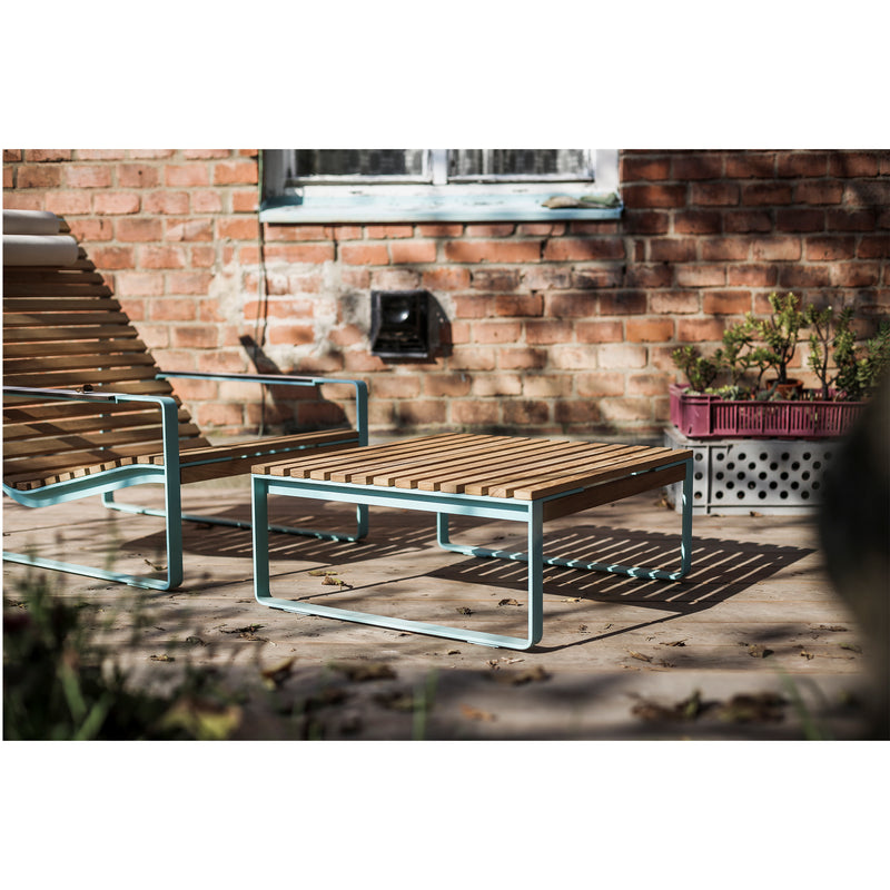 A stylish patio scene features the Egoe Preva Ottoman, with wooden slats and light blue metal frames, set against a sunlit red brick wall. This high-quality furniture enhances the atmosphere alongside a planter box and scattered leaves.