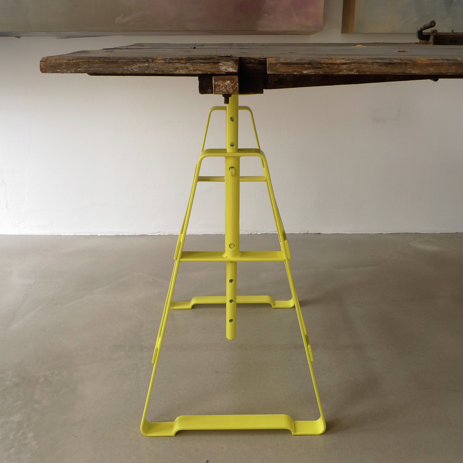 The Lackaffe Trestle from Atelier Haussmann features a wooden tabletop on a height-adjustable, powder-coated steel frame, boasting a minimalist geometric design. It stands on a smooth beige floor against a white wall.