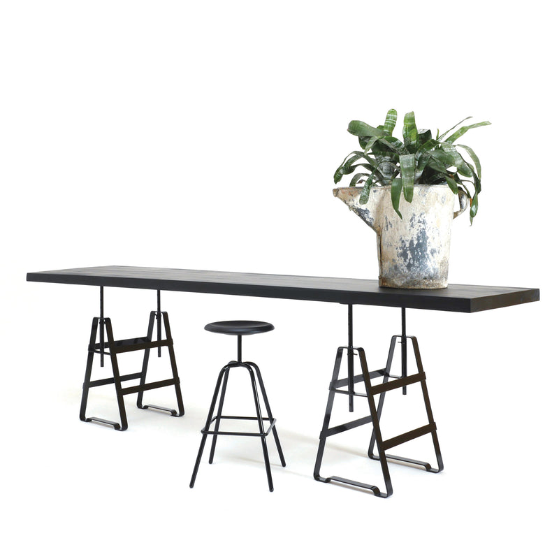 A minimalist setup includes a long dark wooden table with height-adjustable Lackaffe Trestle legs by Atelier Haussmann. A black round stool and a large pot of green leafy plants enhance the scene against a plain white background.