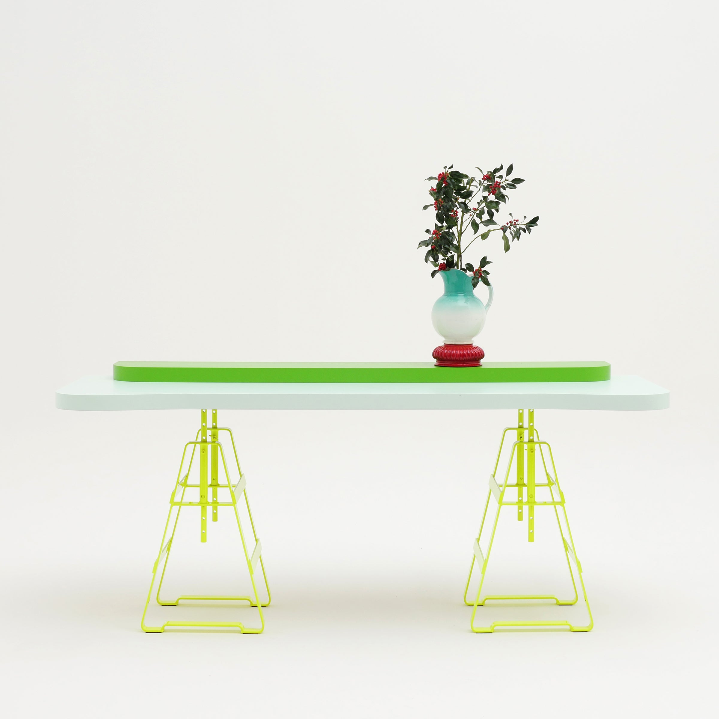The Lackaffe Trestle by Atelier Haussmann showcases a pale green top and powder-coated steel, yellow wireframe legs, with a two-toned blue and red vase holding greenery and red berries, all set against a plain white background.