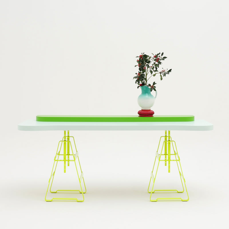 The Lackaffe Trestle by Atelier Haussmann showcases a pale green top and powder-coated steel, yellow wireframe legs, with a two-toned blue and red vase holding greenery and red berries, all set against a plain white background.