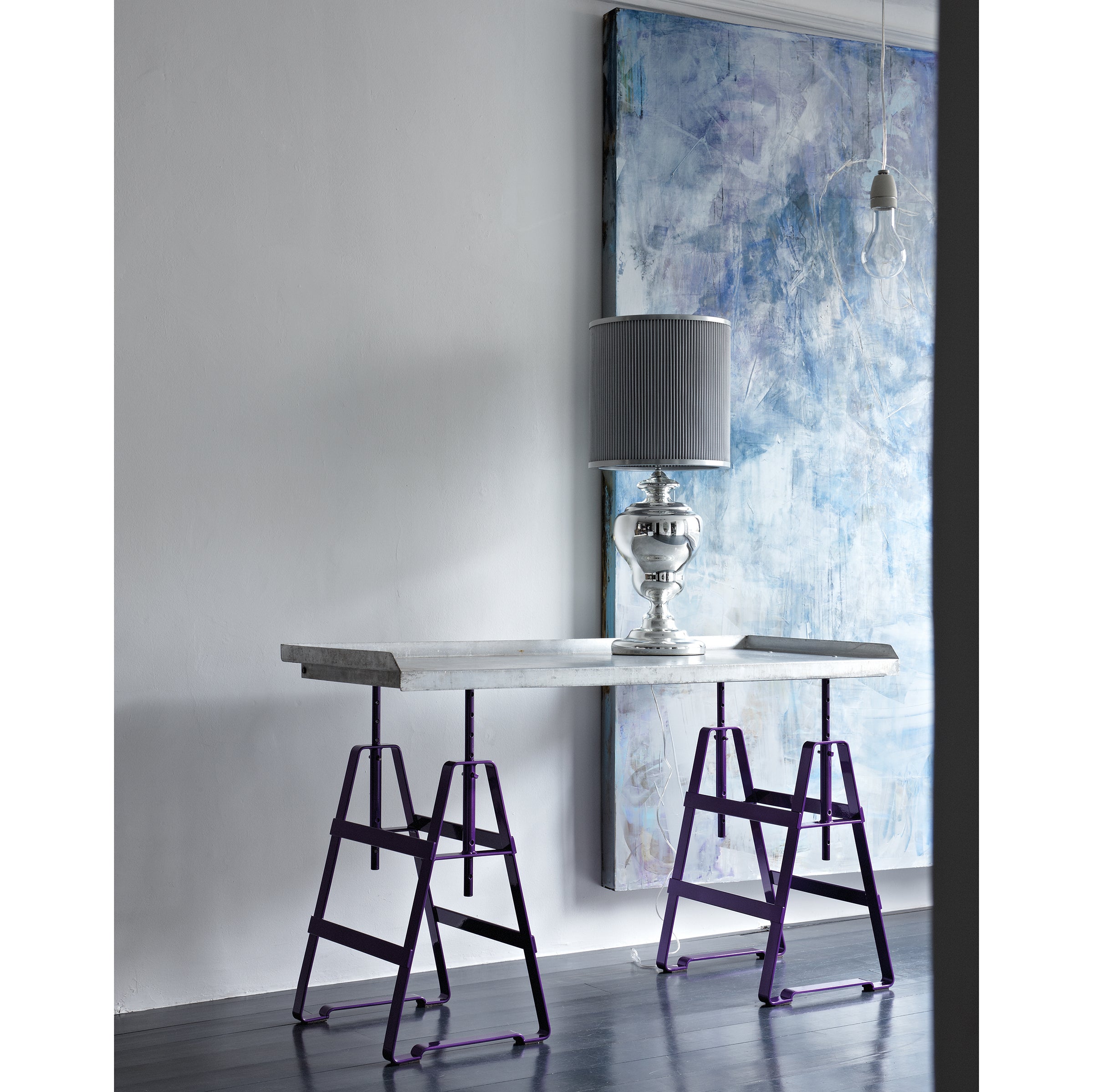 A minimalist room showcases a rectangular table with purple Lackaffe Trestle legs by Atelier Haussmann, featuring a gray marble top. A large lamp with a gray pleated shade rests on the table. A blue abstract painting by Berlin craft producers leans against the white wall, and a bare bulb hangs overhead.
