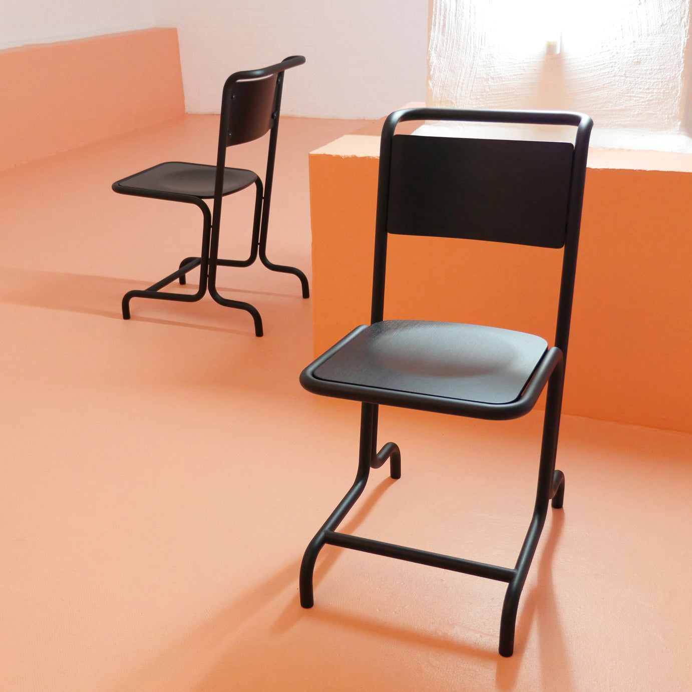 Two László Chairs by Atelier Haussmann, featuring a modern black cantilever design, sit on a peach floor. Positioned near the sunlit window, one chairs sleek lines and comfort are highlighted. The space is bright and airy with soft shadows from the curved steel pipe frames on the floor.