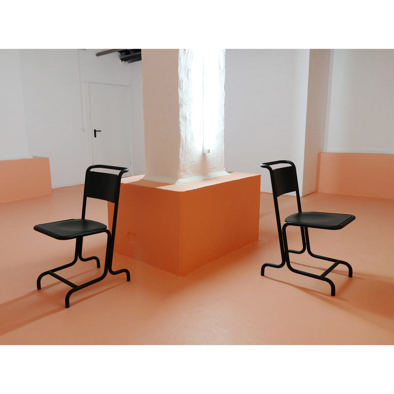 Two László Chairs from Atelier Haussmann face each other on an orange floor in a minimalist room. A large white column stands between them against peach-colored walls, creating a modern atmosphere with clean lines and a bright, uncluttered aesthetic that enhances seating comfort.