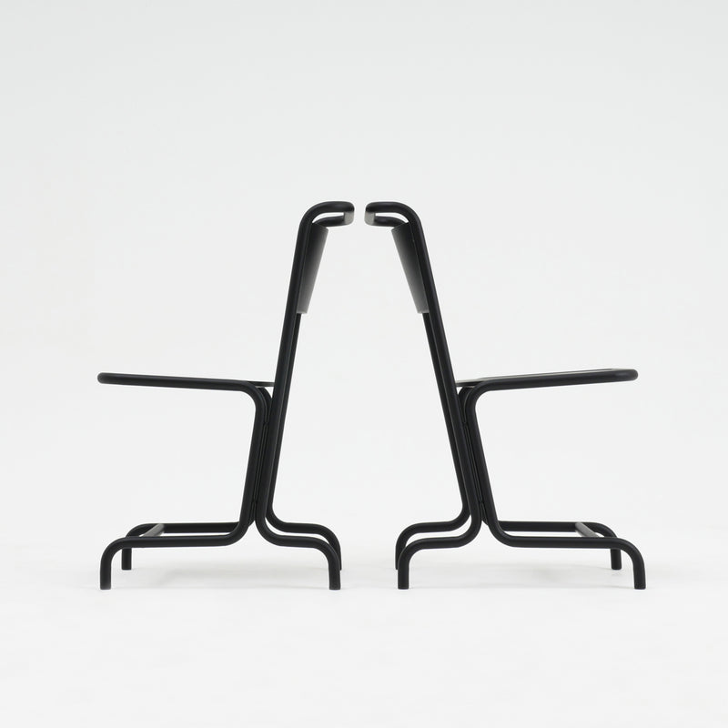 Two identical László Chairs by Atelier Haussmann, crafted from curved steel pipe, are positioned back-to-back on a white background, showcasing seating comfort with their symmetrical and minimalist black metal design.