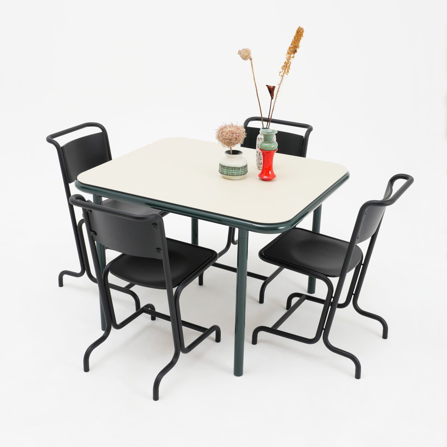 A square table with a light top and dark green legs is surrounded by four Atelier Haussmann László Chairs for comfort. Decorative vases with dried flowers adorn the table, all set against a plain white backdrop.