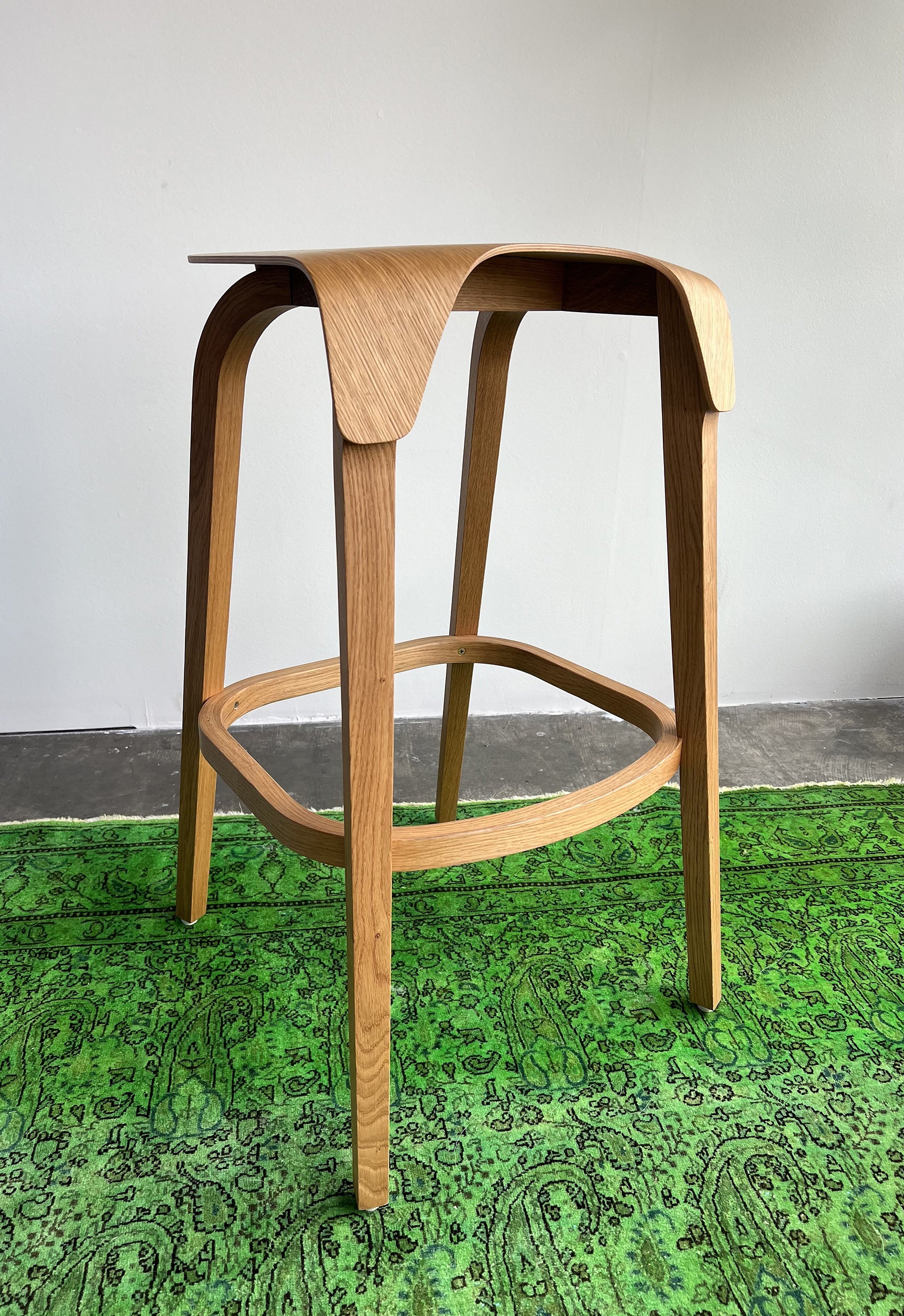 The Barstool Leaf by TON, with its curved, ergonomic design, sits elegantly on a vibrant green rug with intricate patterns. A plain light gray wall in the background complements the stools natural elegance.