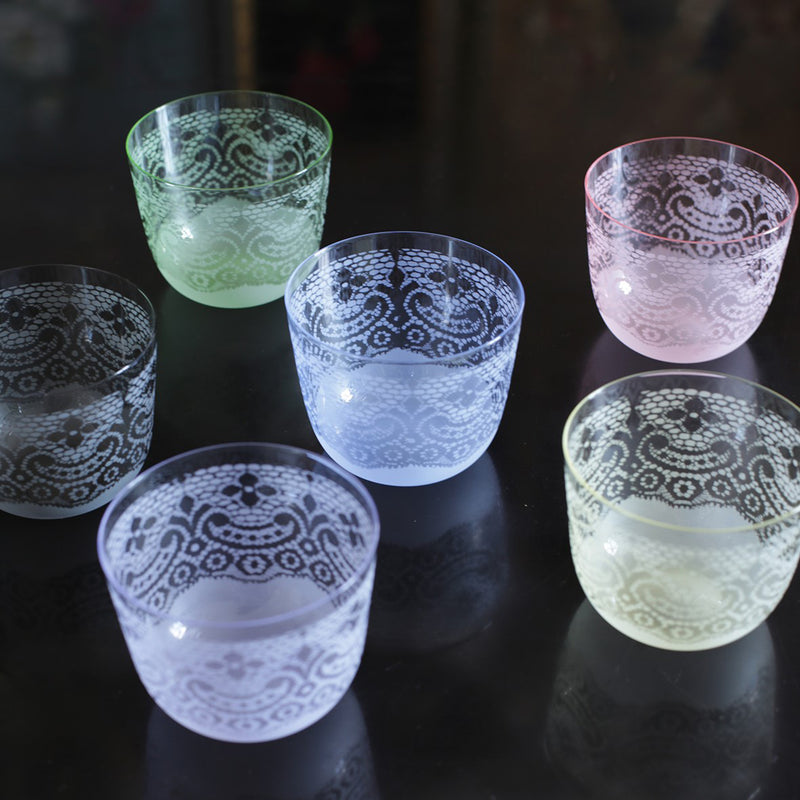 The Alpha Water Tumblers by Lobmeyr come in six translucent pastel shades—green, blue, pink, purple, white, and yellow—and feature intricate floral patterns inspired by Susanne Bisovskys Viennese Chic. They are elegantly displayed on a reflective black surface.