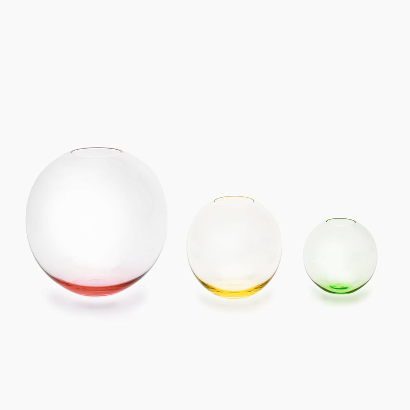 Three Lobmeyr BV60 Flower Vases, crafted from exquisite mouth-blown muslin glass, are arranged from largest to smallest against a white backdrop. The largest has a pink tint at the base, the middle gleams yellow, and the smallest glows green.