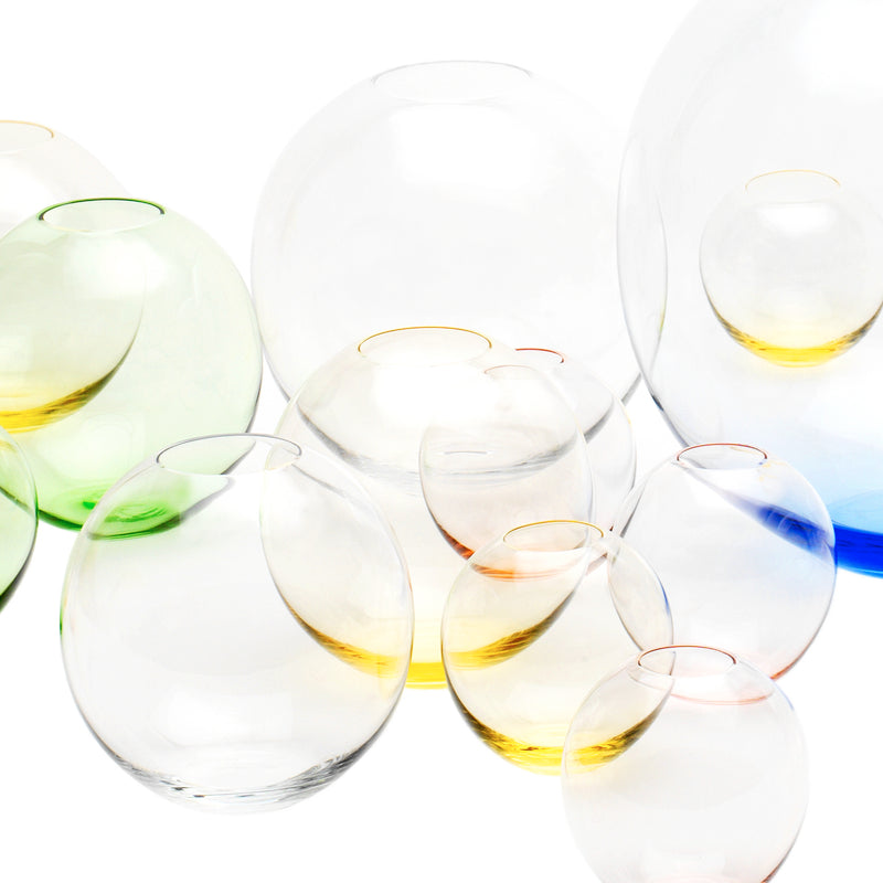 The BV60 Flower Vase by Lobmeyr features mouth-blown transparent glass spheres in various sizes that overlap gracefully. Reminiscent of Stefan Rath’s style, these vases showcase subtle hues of green, yellow, blue, and orange on a pristine white background.