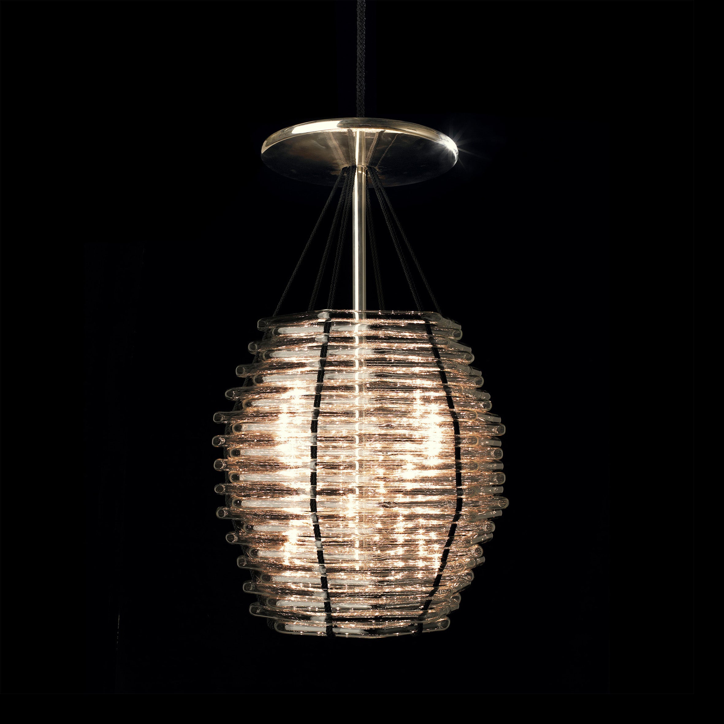 The Basket Chandelier - Sale by Lobmeyr is a modern cylindrical pendant light with stacked glass rings and metal rods. Suspended from a ceiling mount with silk cords, it emits a warm glow against a dark background.