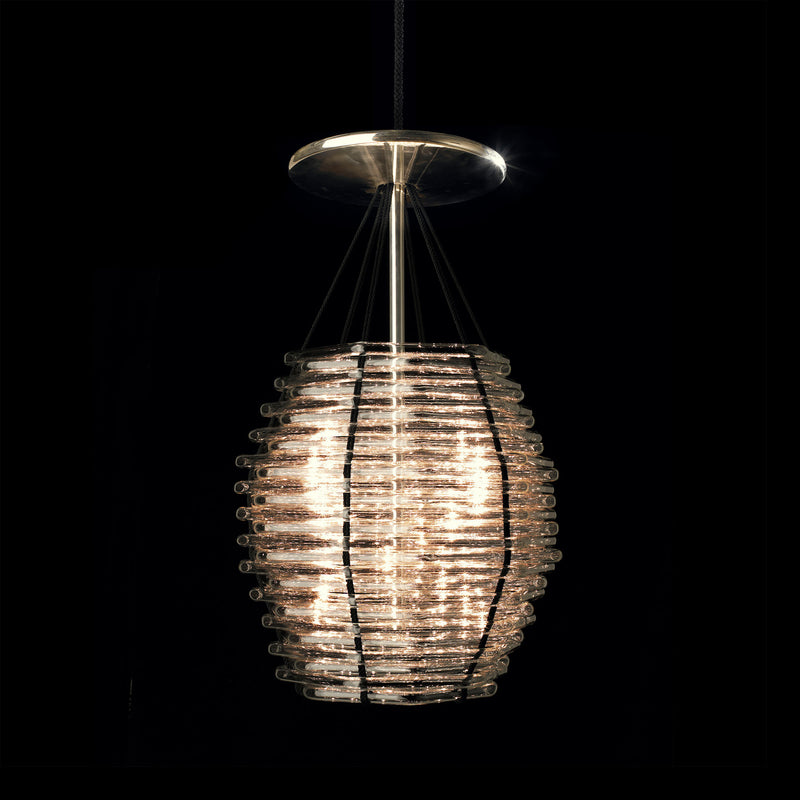 The Basket Chandelier - Sale by Lobmeyr is a modern cylindrical pendant light with stacked glass rings and metal rods. Suspended from a ceiling mount with silk cords, it emits a warm glow against a dark background.